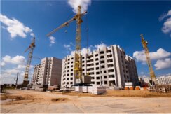 A large construction site with three tall cranes and partially-built concrete apartment buildings under a blue sky with white clouds.